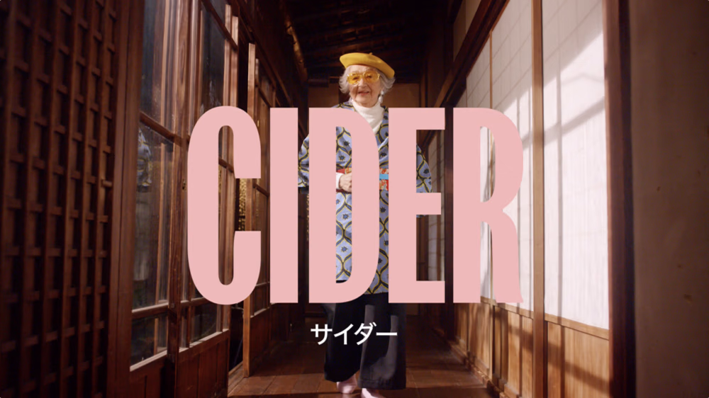 An older person with a cheerful expression walks down a corridor, wearing a bright yellow hat, a colorful patterned jacket, and dark pants. The word "CIDER" in large pink letters overlays the image, with Japanese text for "cider" below it.