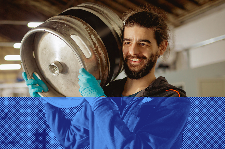 A man with long hair and a beard smiles as he carries a large metal keg on his shoulder. He is wearing a blue hoodie and blue gloves, and is in an industrial or brewery setting. A blue graphic overlay is present at the bottom of the image.