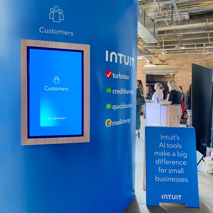 A blue, cylindrical display at an event showcases a touchscreen labeled "Customers" with a sign highlighting Intuit's services, including TurboTax, Credit Karma, QuickBooks, and Mailchimp. Another sign reads, "Intuit’s AI tools make a big difference for small businesses.