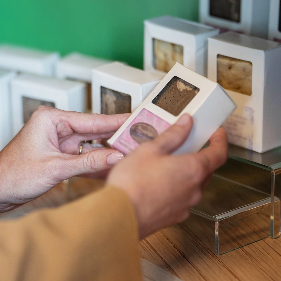 A person is holding a box containing two pieces of soap with visible windows. More similar boxes are displayed in the background on a wooden surface with a green wall. The person appears to be examining or selecting a box.