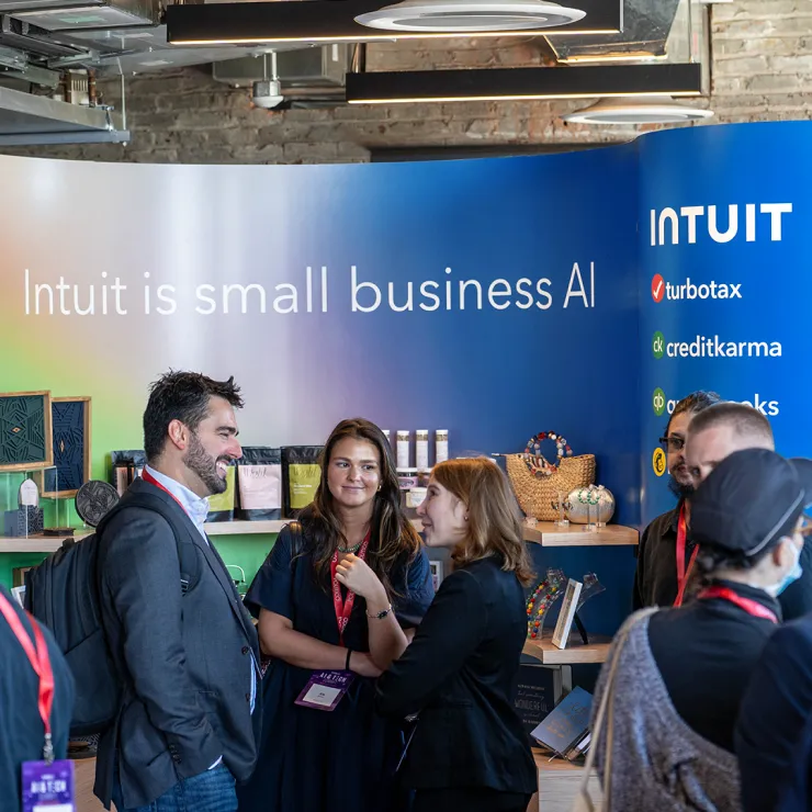 A group of people are having a conversation at an exhibit booth with a colorful background displaying the text "Intuit is small business AI." Logos for TurboTax, Credit Karma, and QuickBooks are visible. Shelves with various items are in the background.