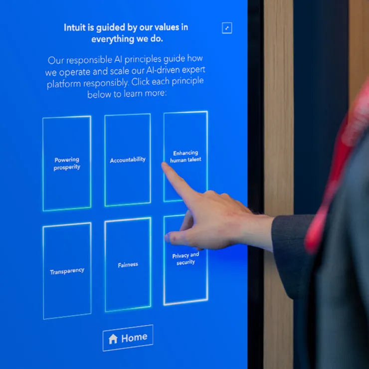 A person is interacting with a touchscreen display that has a message about Intuit's values. The display shows six principles: "Powering prosperity," "Accountability," "Enhancing human talent," "Transparency," "Fairness," and "Privacy and security.