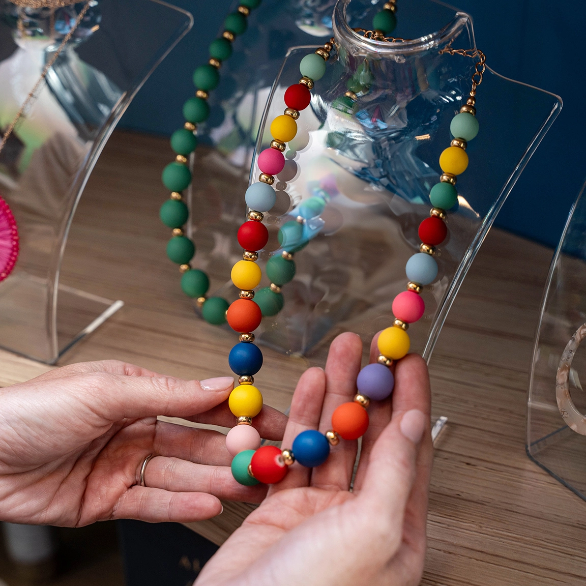 A pair of hands is holding a colorful beaded necklace. The necklace, made up of large, round beads in various bright colors, is displayed on a clear bust. In the background, there are other beaded necklaces in different colors on similar clear busts.