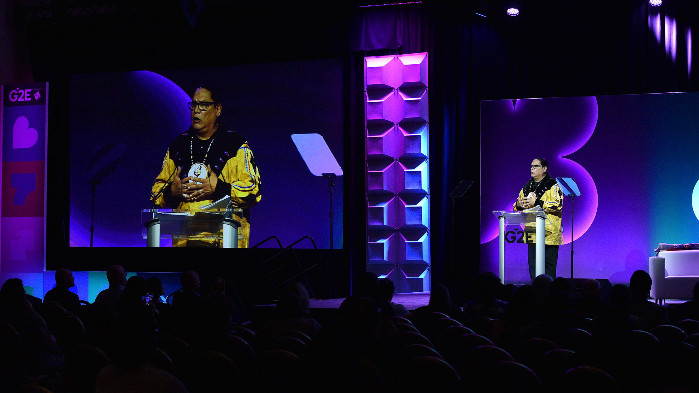 A speaker stands at one of two podiums on a stage, addressing an audience at G2E. The speaker appears in a close-up on a large screen to the left. The background features purple and blue lighting with geometric patterns. The audience sits in dim lighting.