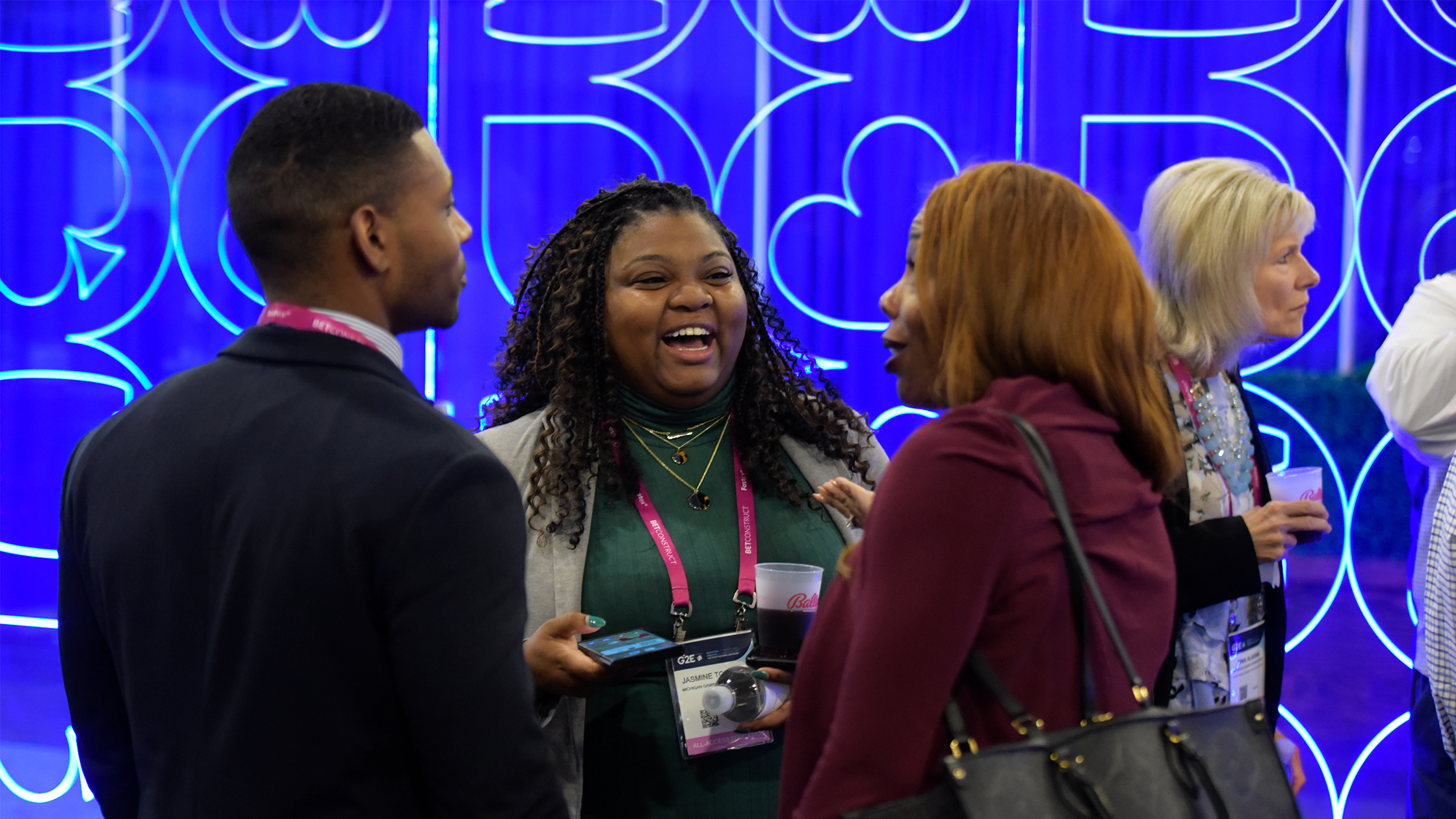 A diverse group of five people interact at a conference, standing in front of a blue display featuring heart and infinity symbols. The woman in the center has curly hair and is laughing, engaging with two others. They wear badges and lanyards, suggesting an event setting.