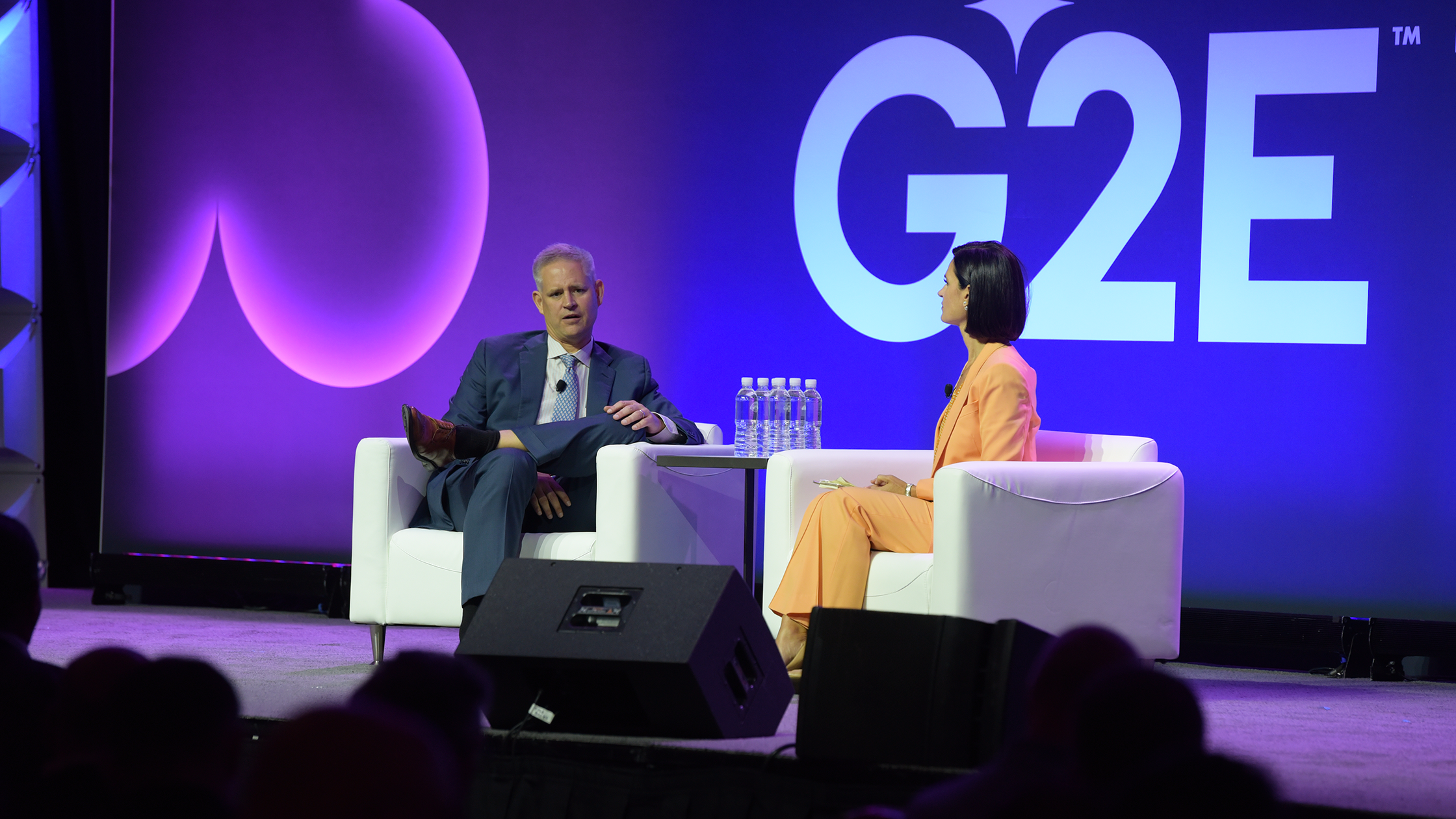 Two people are seated on white chairs on a stage in front of a backdrop displaying "G2E" in large text. The person on the left is speaking while the person on the right listens attentively. There are three water bottles on a table between them. The lighting is predominantly purple.