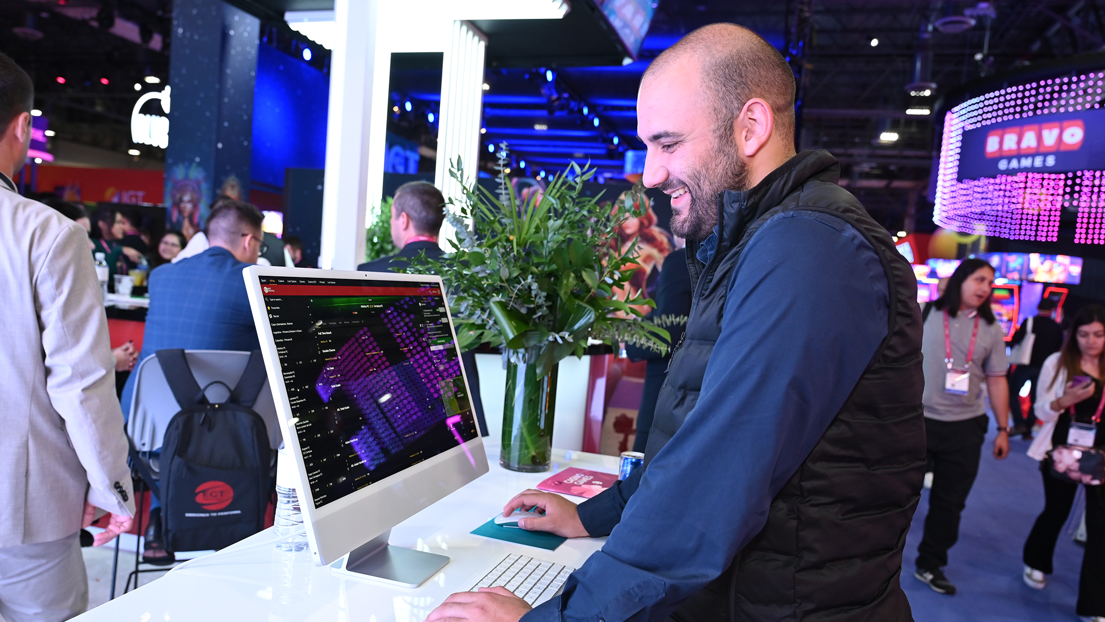 A man stands at a high counter using a desktop computer, smiling while interacting with the screen. The setting appears to be a lively event or trade show with colorful booths, displays, and other attendees visible in the background. A vase of greenery is on the counter.