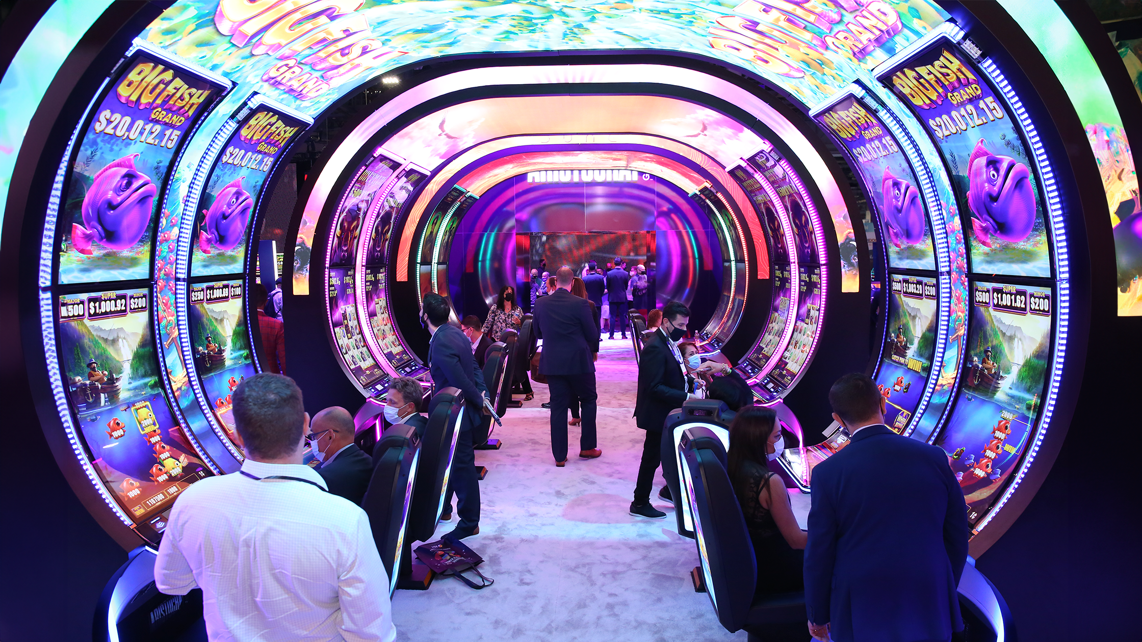 A vibrant casino floor featuring a colorful, illuminated tunnel-like structure filled with numerous slot machines. People are seated at the machines, engrossed in playing, while others stand and walk around. The environment is lively and bustling with activity.