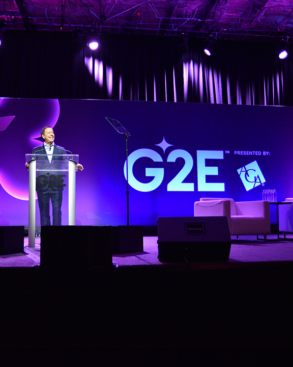 A person stands behind a podium on a stage speaking at the Global Gaming Expo (G2E), presented by the American Gaming Association (AGA). The stage is lit with purple lighting, and there is a white chair and a small table in the background.