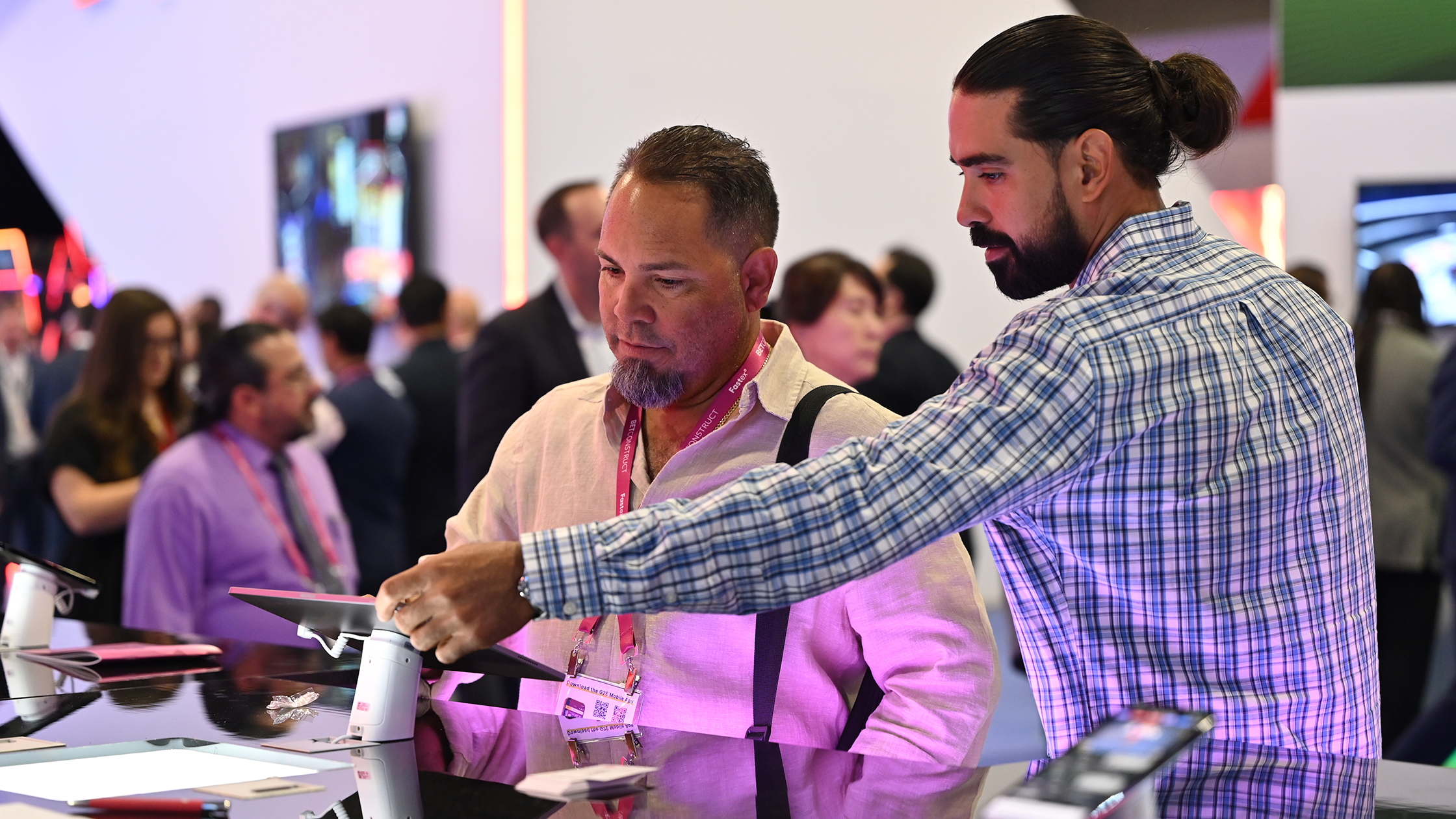 Two men at an event are interacting with a tablet device on a table. One man, in a light shirt with suspenders, examines the device, while the other, in a blue plaid shirt, points something out on the screen. People in formal attire are gathered in the background.