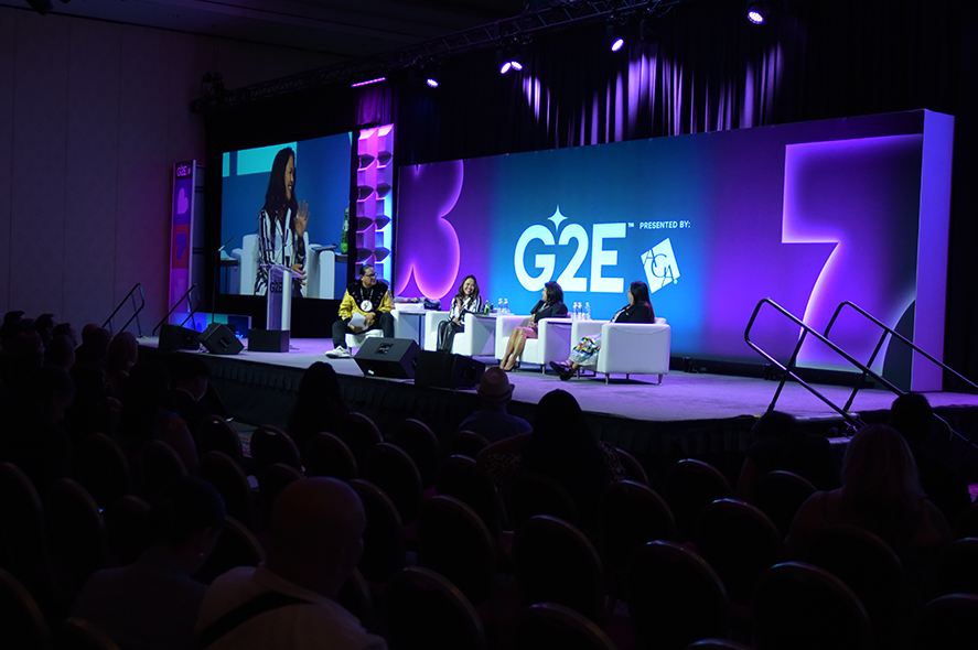 A conference panel discussion with four speakers seated on a stage with purple lighting. The stage backdrop displays "G2E" in large letters. A screen to the left shows a live feed of one of the speakers. Attendees are seated and facing the stage.