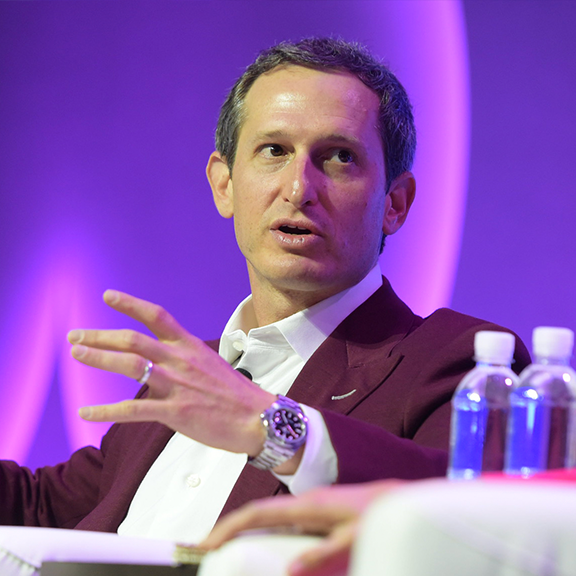 A man in a maroon blazer and white shirt is speaking, with his hand raised and gesturing. He is seated and looking to his right. Two bottles of water are placed on a table in the foreground. The background is lit with a purple hue.