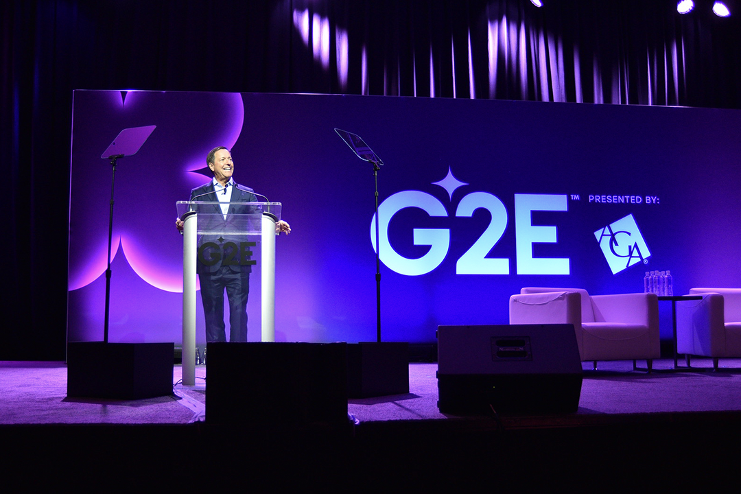 A person stands at a transparent podium on a stage, delivering a speech in front of a large "G2E" sign. The stage is lit with purple lighting, and empty white chairs are positioned to the right. The backdrop includes the text "Presented by:" followed by a logo.