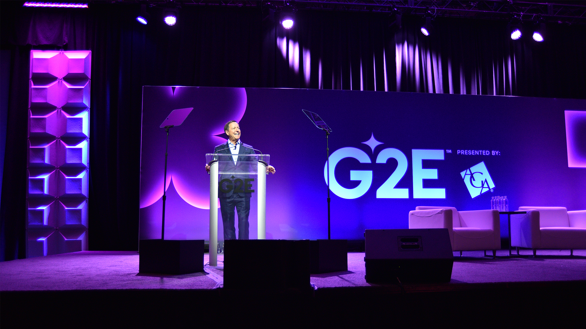 A person is standing at a podium on a stage, speaking at an event with the backdrop displaying "G2E" and "Presented by". The stage is lit with purple lighting, and two empty white chairs are visible to the right of the speaker.