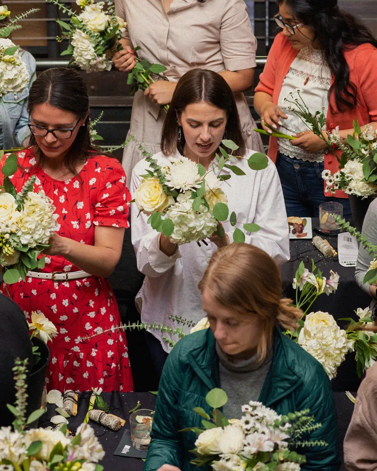 People are gathered around tables arranging flower bouquets. The group includes mostly women, who are focused on creating their floral arrangements with various white and yellow flowers and green foliage. The setting appears to be an indoor workshop or class.
