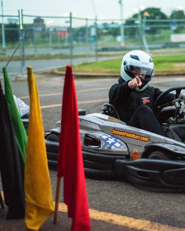 A person wearing a helmet and racing attire sits in a go-kart on a track, pointing towards the camera. Colored flags, including green, yellow, and red, are standing in the foreground. The background shows a fenced area and blurred outdoor scenery.