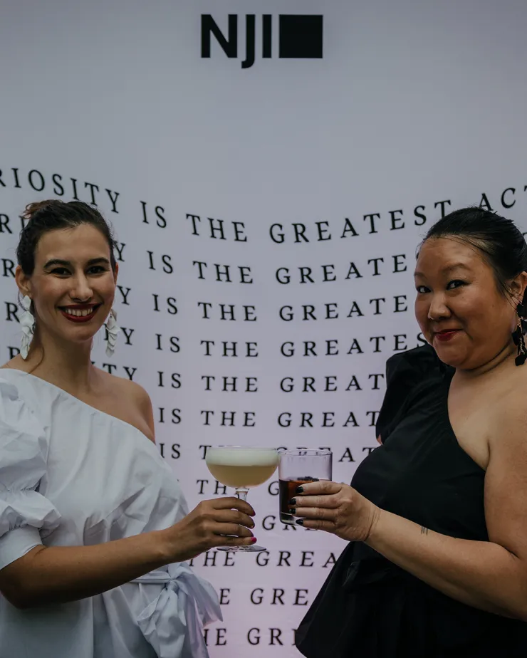 Two women smile and clink glasses in front of a backdrop featuring the repeated phrase "CURIOSITY IS THE GREATEST ACT." One woman wears a white, one-shoulder dress, and the other wears a black, off-shoulder dress. The logo "NJII" is seen at the top of the backdrop.