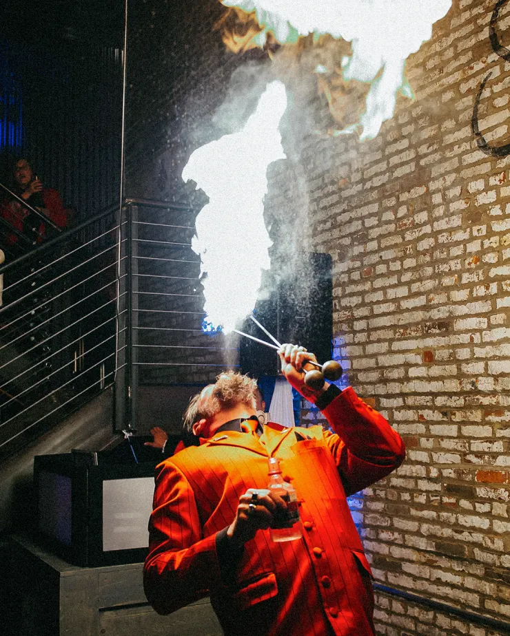 A performer in a vibrant red suit breathes fire towards the ceiling in an indoor setting with brick walls. He holds a fire-breathing torch in one hand and a container in the other. Onlookers are visible in the background, standing near a metal staircase.