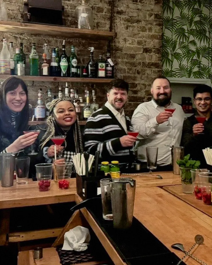A group of five people smiling and holding cocktails in a bar. They are standing behind a bar counter with various bottles and bar equipment around. The background features a brick wall with shelves of bottles and a section with green leafy wallpaper.