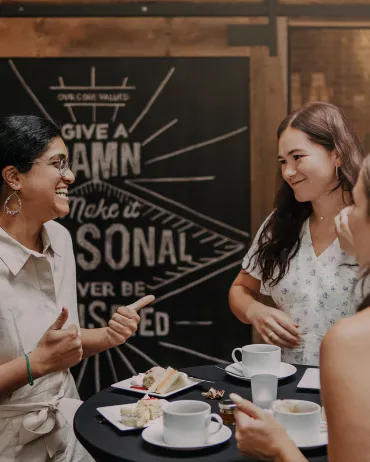 Three women are standing and engaging in conversation around a small table with coffee cups and plates of pastries. One woman is gesturing with her hands while the two others are listening and smiling. The background features a decorative wall with text.