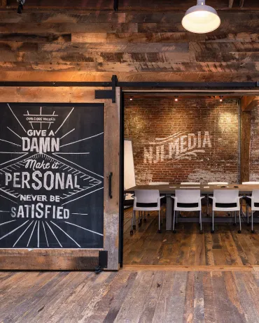 A rustic meeting room is visible through a large wooden sliding door. The door has a motivational quote on it. Inside, brick walls, a table, and chairs are illuminated by warm light, and "WJ MEDIA" is painted on the wall. The room has a cozy, wood-paneled floor.