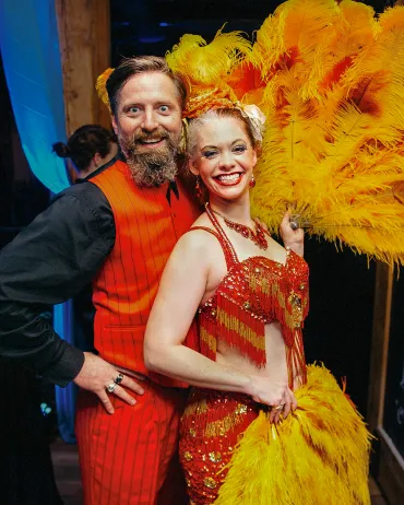A man with a beard wearing a red vest stands next to a smiling woman dressed in an elaborate red and gold fringed costume. She holds a large yellow feather fan, and both appear to be enjoying a festive event.