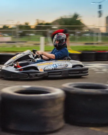 A person wearing a helmet and protective gear drives a go-kart on an outdoor track surrounded by tires. The go-kart and driver are captured in motion, with blurred background elements indicating speed. The setting is during the evening with a soft orange hue in the sky.