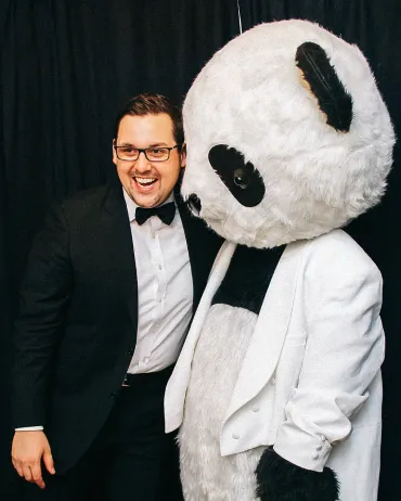 A man in a black tuxedo with a bow tie smiles and poses next to a person wearing a large panda costume with a white jacket and black pants. They are standing in front of a black curtain backdrop.