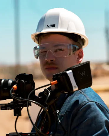A person wearing a white hard hat, safety goggles, and a dark blue jacket is standing outdoors. They are holding a piece of equipment with several wires and a screen attached, possibly a camera or surveying device. The sky is clear and the background is lightly blurred.