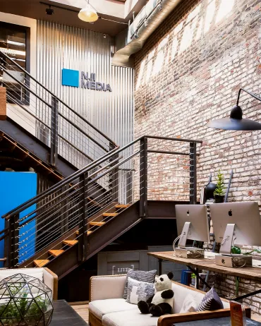 Modern office space with exposed brick walls, metal stairs, and a large "NU MEDIA" logo on the wall. The area features desks with computers, a cozy seating area with cushions, a stuffed panda, and a geometric terrarium on a wooden table. Industrial lighting overhead.