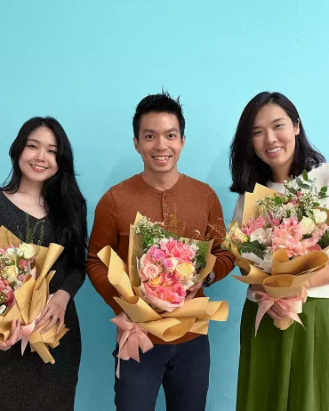 Three people are standing in front of a light blue wall, each holding a bouquet of flowers wrapped in brown paper. The person in the middle is wearing a rust-colored shirt, the person on the left a black outfit, and the person on the right a white top with a green skirt. They are all smiling at the camera.