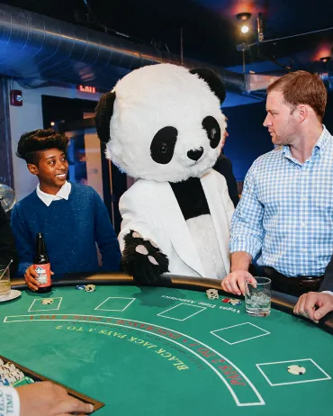 Two people are playing blackjack at a casino table, with a person dressed in a panda costume standing between them. The person on the left is holding a drink and smiling, while the person on the right, in a checkered shirt, is looking at the panda.