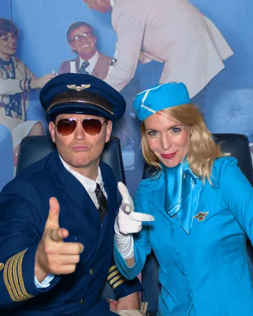 A person dressed as a pilot and another person dressed as a flight attendant, both in vintage-style uniforms, pose playfully. The pilot wears sunglasses and points finger-gun gestures, while the flight attendant smiles and mimics the gesture. A blue backdrop is behind them.