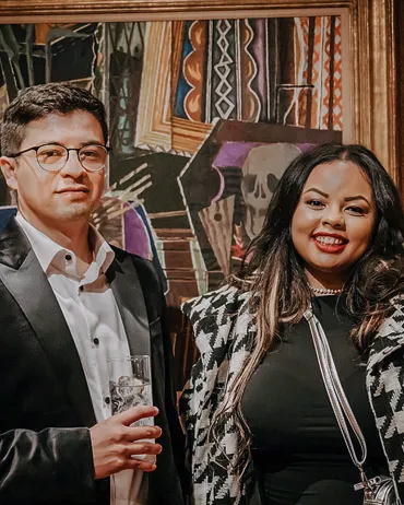 A man and a woman stand in front of a colorful, abstract painting. The man wears glasses, a black suit jacket, and a white shirt, holding a drink. The woman has long dark hair, smiles, and wears a black and white patterned jacket over a black top.