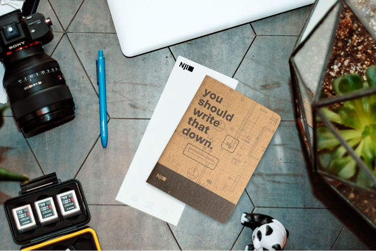 A flat lay image features a wooden table with a camera, lens cap, several SD cards, a pen, a closed laptop, a terrarium with plants, and a notebook with a text that reads "you should write that down." The table has hexagonal patterns on its surface.