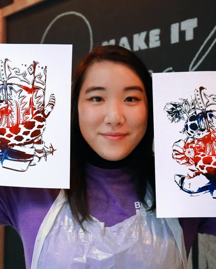 A woman is holding up two pieces of art, one in each hand. The artwork features abstract designs with a mix of red, blue, and black colors. She is wearing a purple top and a transparent apron, standing in front of a blackboard with partial text visible.