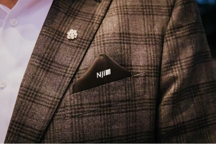 Close-up of a person wearing a plaid suit jacket with a white shirt underneath. The suit jacket has a flower-shaped pin on the lapel and a pocket square with the initials "NJI" embroidered on it in white. The background is blurred.