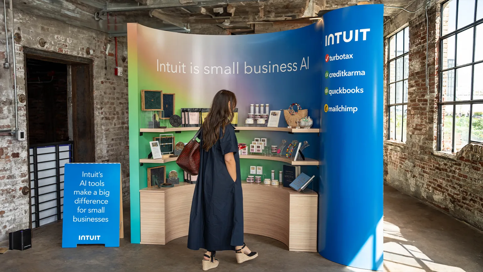 A woman in a black dress stands in front of a curved display booth showcasing AI tools for small businesses. The booth features labels for various Intuit products, including Turbotax, CreditKarma, QuickBooks, and Mailchimp, with shelves of products and decor.