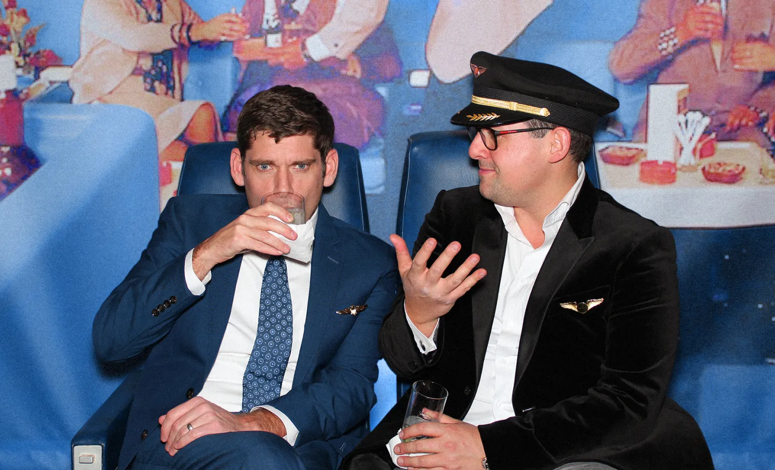 Two men are sitting side by side. The man on the left is wearing a blue suit and tie, holding a drink. The man on the right is dressed in a pilot's uniform, also holding a drink, and appears to be talking. There's a colorful background behind them with people and tables.