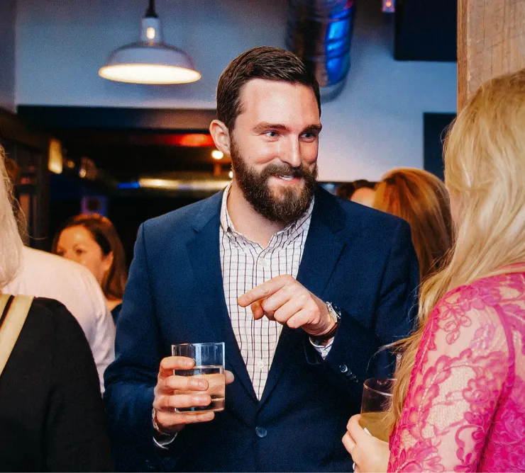 A man with a beard, dressed in a blue suit and checkered shirt, is holding a drink and smiling while engaging in conversation with a woman in a pink lace dress. The setting appears to be a social gathering or party with other people in the background.