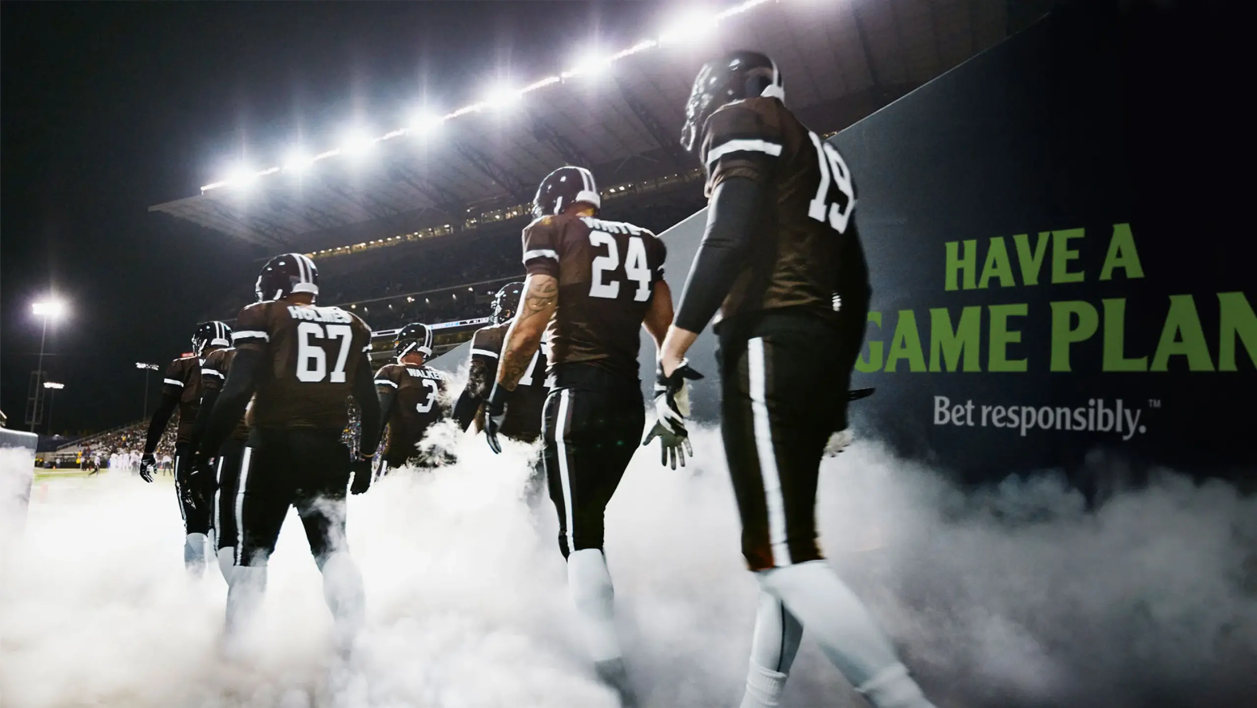A football team wearing black jerseys with white pants walks through smoke onto the field. The stadium lights are bright in the background. A large sign reads, "Have a game plan. Bet responsibly." The players' jersey numbers visible are 67, 24, and 19.
