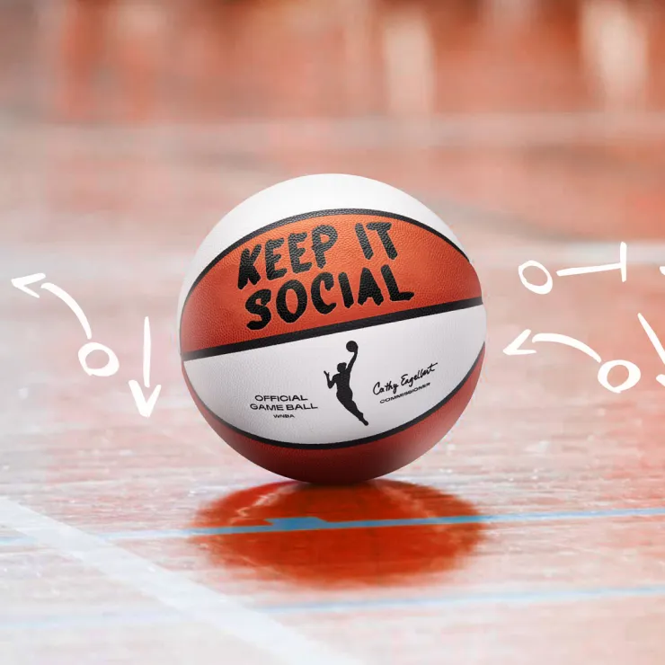 A basketball with "KEEP IT SOCIAL" written on it is placed on an indoor court. The ball, featuring a figure of a female player in black, is encircled by white doodle arrows pointing towards it. The text "Official Game Ball" and "Cathy Engelbert" is also visible on the ball.