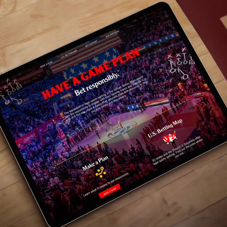A tablet showing a sports betting website with a basketball game background. The text "HAVE A GAME PLAN" and "Bet responsibly." is prominent. Options "Make a Plan" and "U.S. Betting Map" are available. The wooden surface beneath the tablet is partially visible.