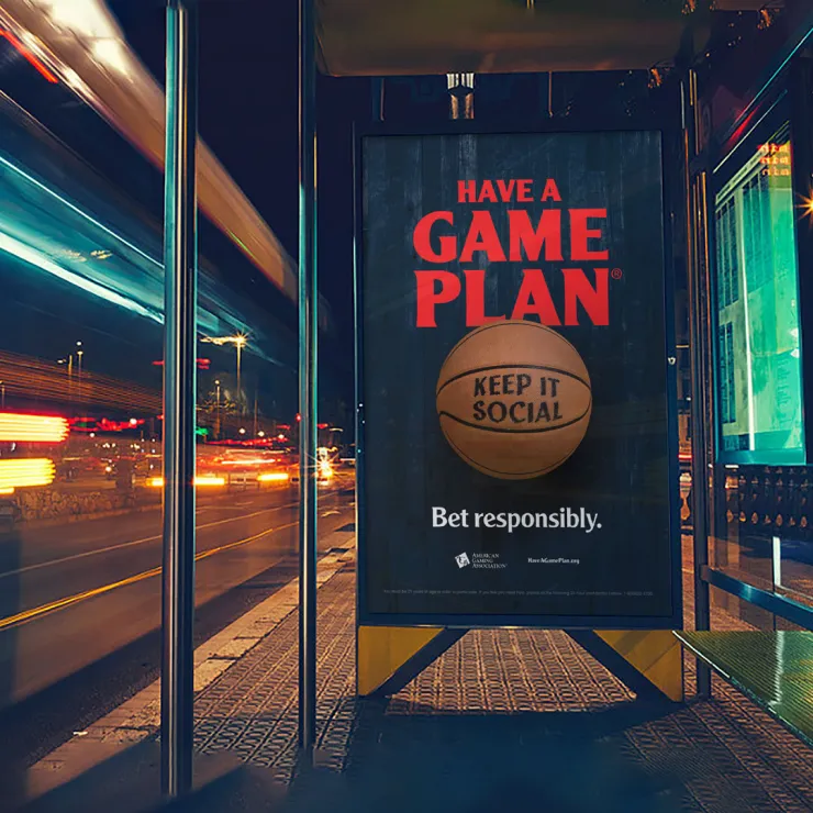 A bus stop advertisement at night shows a basketball with the text "KEEP IT SOCIAL" on it. Above the ball, in bold red letters, it reads "HAVE A GAME PLAN." Below the ball, it says "Bet responsibly." The scene is illuminated by streetlights and moving lights from traffic.