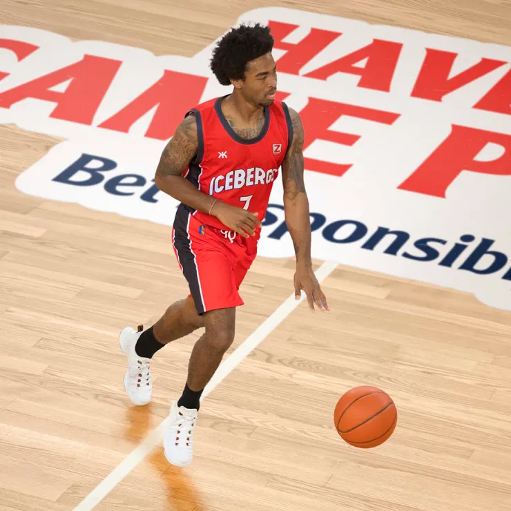 A basketball player in a red "Iceberg" uniform and white sneakers dribbles the ball on a wooden court. The player has a tattooed right arm and sports a headband. The court features a large red and white advertisement in the background.