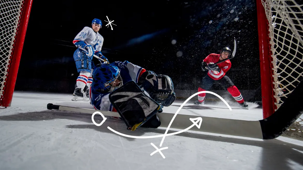 A hockey player in a red jersey shoots the puck towards the goal as the goalie in a blue jersey dives to make a save. Another player in a white jersey watches from the background. Hand-drawn play strategy lines overlay the image, indicating the action.