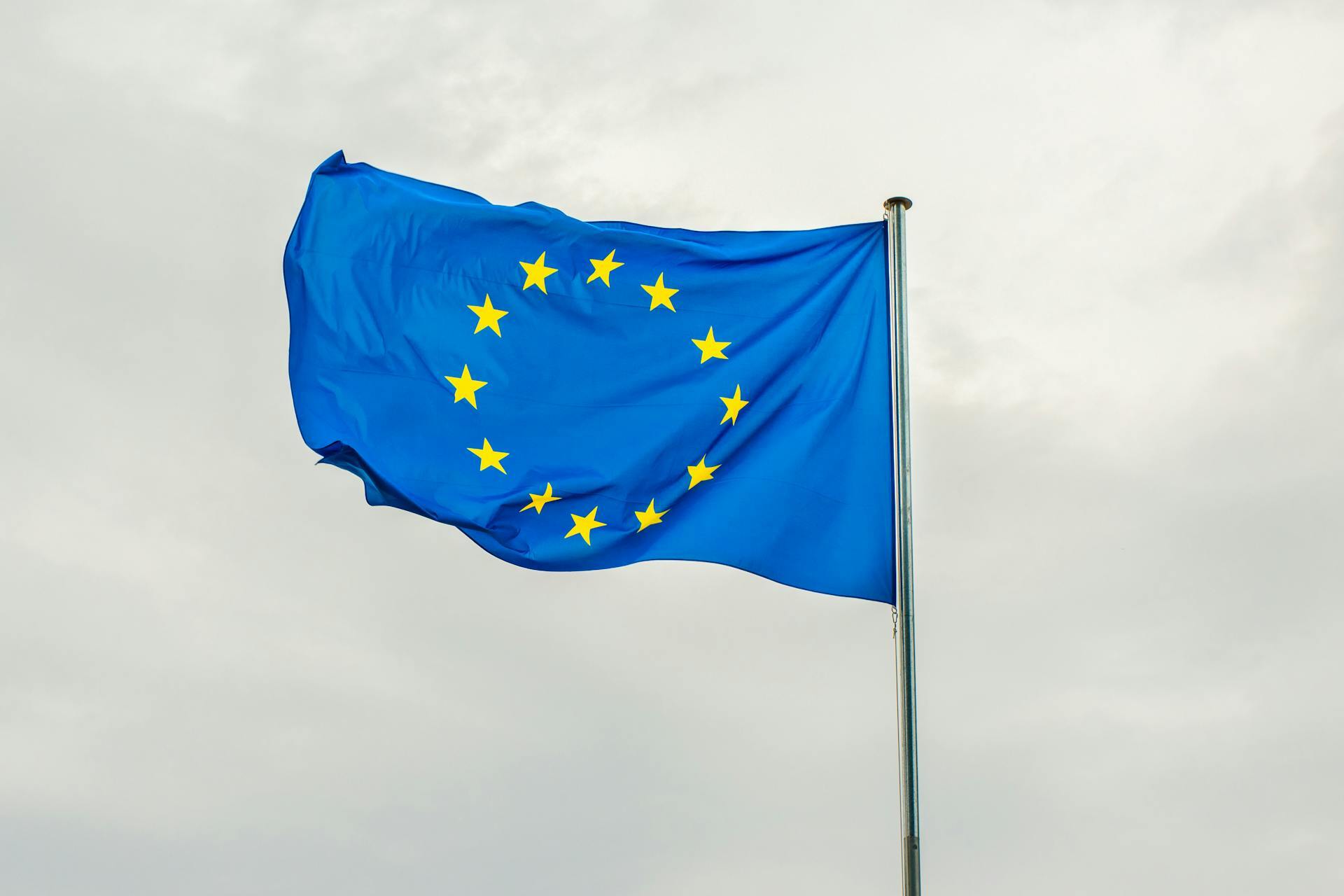 The image shows the flag of the European Union. It is a blue flag with twelve gold stars arranged in a circle. The flag is waving in the wind against a cloudy sky, and is attached to a long vertical pole.