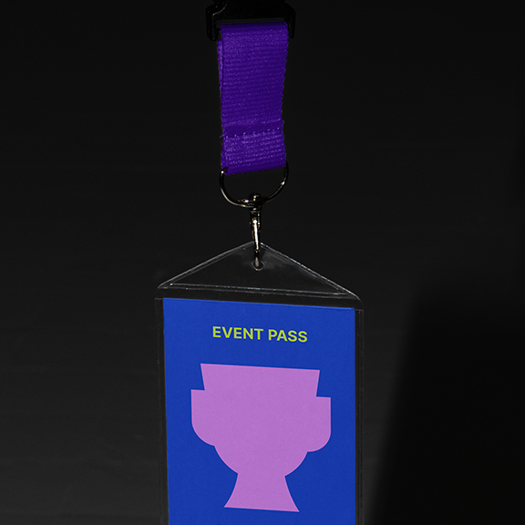 A purple lanyard holds a rectangular event pass with a blue background. The pass features the words "EVENT PASS" in yellow and a silhouette of a trophy or cup in pink. The lanyard is attached to the pass with a metal clip.