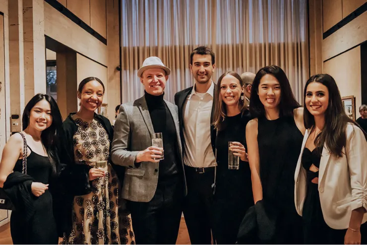 A group of seven people, dressed in semi-formal attire, stand together in a warmly lit room. They are smiling and holding drinks, suggesting a social gathering or event. The background features tall curtains and contemporary decor.