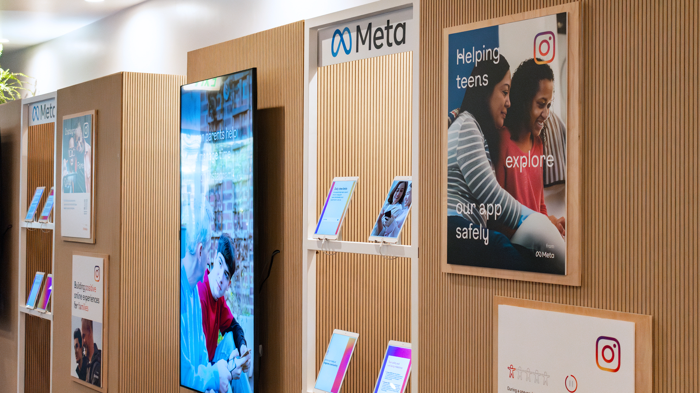 A display area with wooden paneling showcasing Meta's initiatives, including a large monitor and posters. One poster reads "Helping teens explore our app safely" with an image of two individuals. Various informative materials are placed on shelves below the posters.